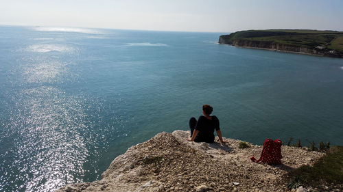 Rear view of woman with backpack sitting on cliff by sea against clear sky