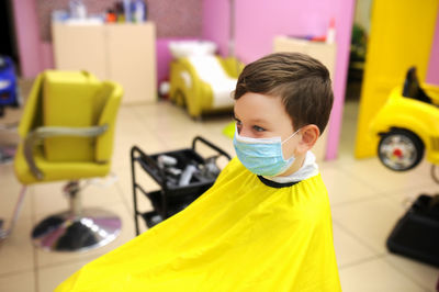 Cute boy wearing mask sitting at barber shop