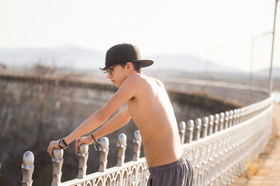 Side view of young man standing on railing against sky