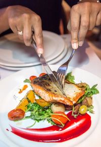 Cropped image of person eating salmon while using table knife and fork