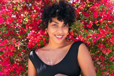 Content young ethnic female with curly hair looking at camera against blossoming shrub on sunny day