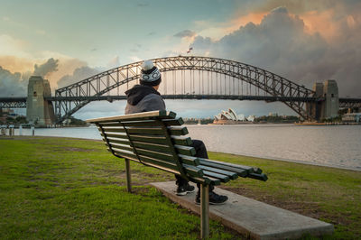 Man sitting on bench by bridge against sky