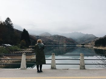 Rear view of woman standing by railing on bridge over lake against sky