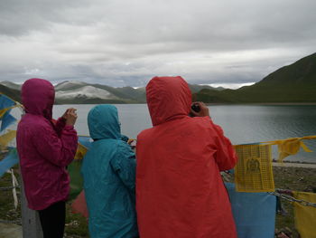 Rear view of women photographing lake and mountains against cloudy sky