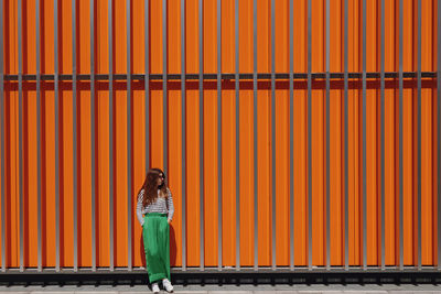 Portrait of stylish woman in geen pants near metall orange wall in urban space