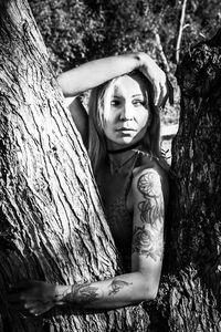 Side view of thoughtful woman with tattoos on hand standing by tree trunk