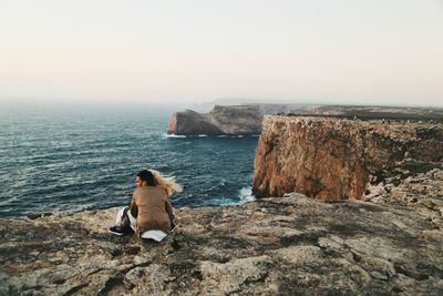 Rear view of man sitting on cliff by sea against clear sky