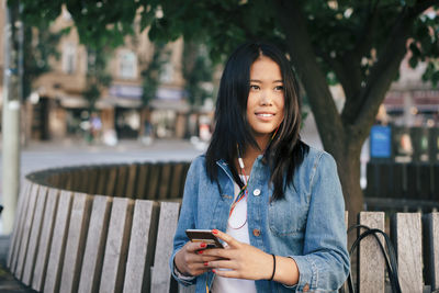 Smiling teenage girl with mobile phone sitting on bench in city