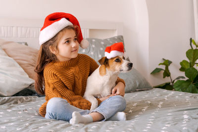 Happy little girl sitting on bed and hugging with dog jack russell in sweater