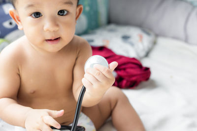 Portrait of cute baby boy holding stethoscope while sitting on bed at home