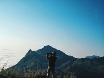 Rear view of man photographing while standing on mountain against clear sky