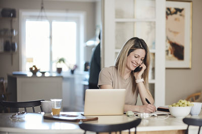 Happy freelancer taking down notes while talking on smart phone at home