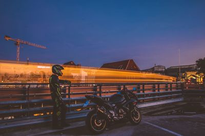 Side view of biker standing by motorcycle against light trails in city at dusk