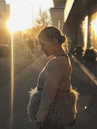 Side view of young woman looking down while standing on city street during sunset