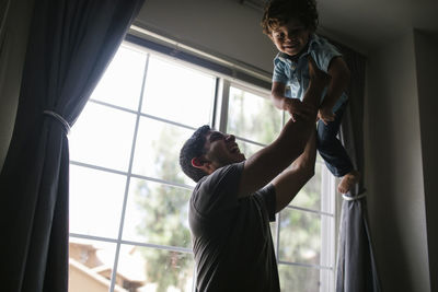 Happy father lifting son while standing by window at home