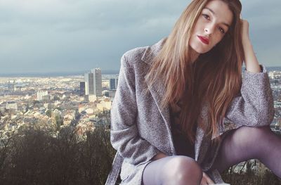Beautiful young woman sitting on cityscape against sky