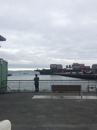 Man standing on railing by sea against sky in city