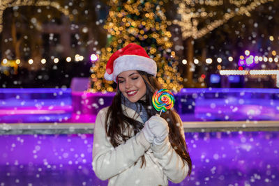 Smiling young woman wearing santa hat holding lollipop in city at night