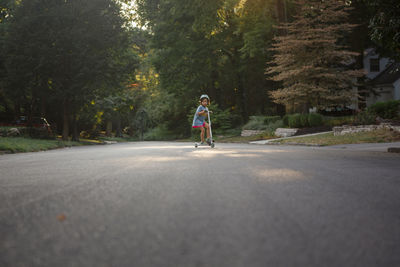 A small child rides on a scooter on a tree-lined street in sunlight