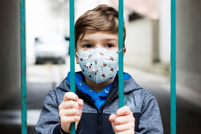 Small boy wearing protective face mask while looking through the gate due to coronavirus pandemic.