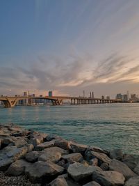 Bridge over sea by buildings against sky during sunset