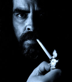 Close-up portrait of man lighting cigarette against black background