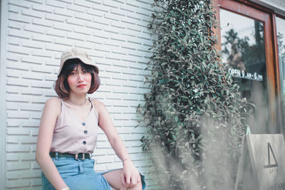 Portrait of young woman wearing hat while sitting against wall