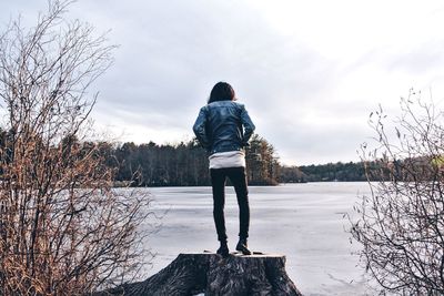 Rear view of woman standing on tree stump by lake against sky