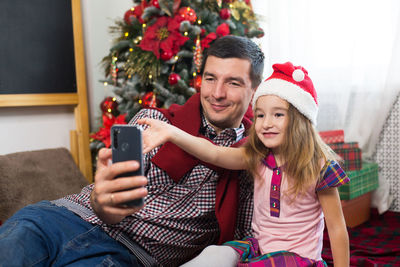 Portrait of smiling young woman using mobile phone while sitting on christmas tree