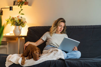 Woman sitting at couch, stroking her dog and working on laptop, surfing web at home in living room