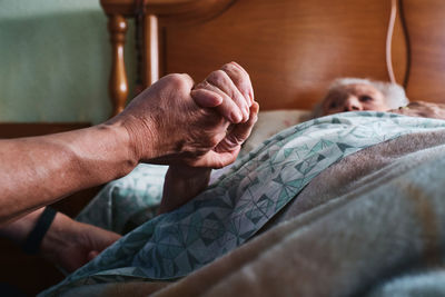 Cropped unrecognizable nurse holding hand of aged female patient with gray hair lying on bed in bedroom at home