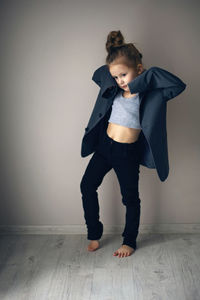 Funny portrait of fashionable girl child in large gray jacket stands against gray wall next  window 