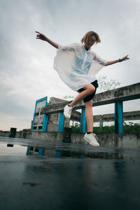 Full length of young woman wearing raincoat while jumping over puddle against sky