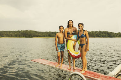 Smiling girl with inflatable ring standing with siblings on diving board at lake