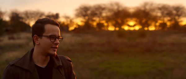 Portrait of young man wearing sunglasses standing on field against sky during sunset