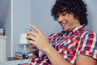 Close-up of smiling man using mobile phone while sitting on bed at home