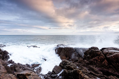 Waves splashing on rocks at shore against sky