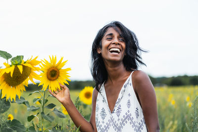 Sincere adult ethnic female looking away on meadow touching blossoming flowers in countryside on blurred background