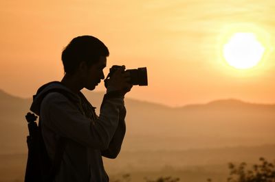 Side view of man photographing with camera against sky during sunset