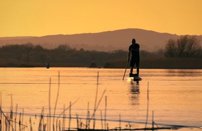 Silhouetted man stand peddle boarding at sunset, river corrib, galway, ireland