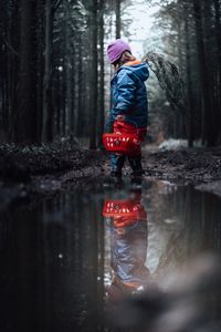 Reflection of girl holding basket in puddle standing against trees in forest