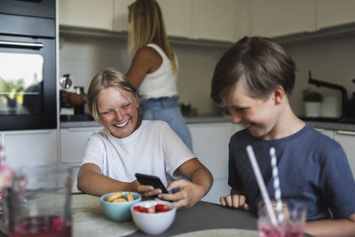 Smiling brothers using smart phone while mother doing kitchen chores in background at home