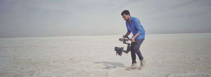 Full length of man filming with camera on gimbal while walking at salt flat against sky