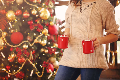 Midsection of woman holding coffee cups against christmas tree
