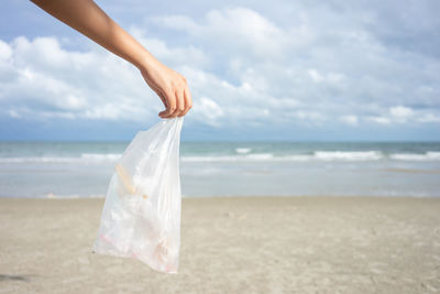 Cropped hand holding plastic bag on beach against sky