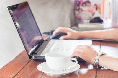 Cropped hands of woman using laptop by coffee cup on table