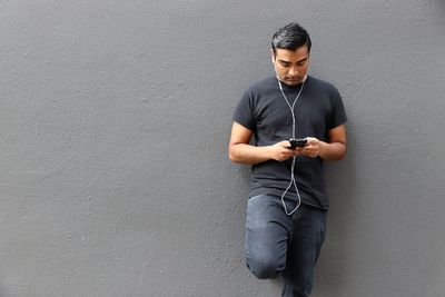 Young man listening music while using mobile phone against wall