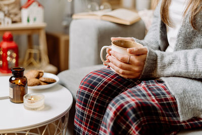 Cozy woman in knitted winter sweater and  pajama drinking hot cocoa, coffee in mug, on couch home.
