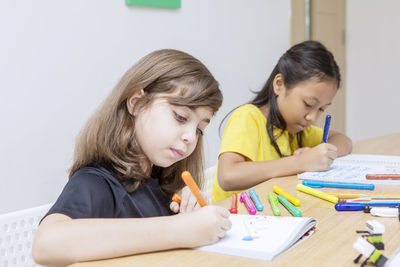 Close-up of girl drawing in book with friend while sitting in classroom at school