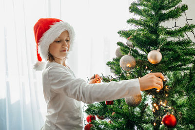 Charming girl in a santa claus hat decorates the christmas tree at home with a garland with lights. 